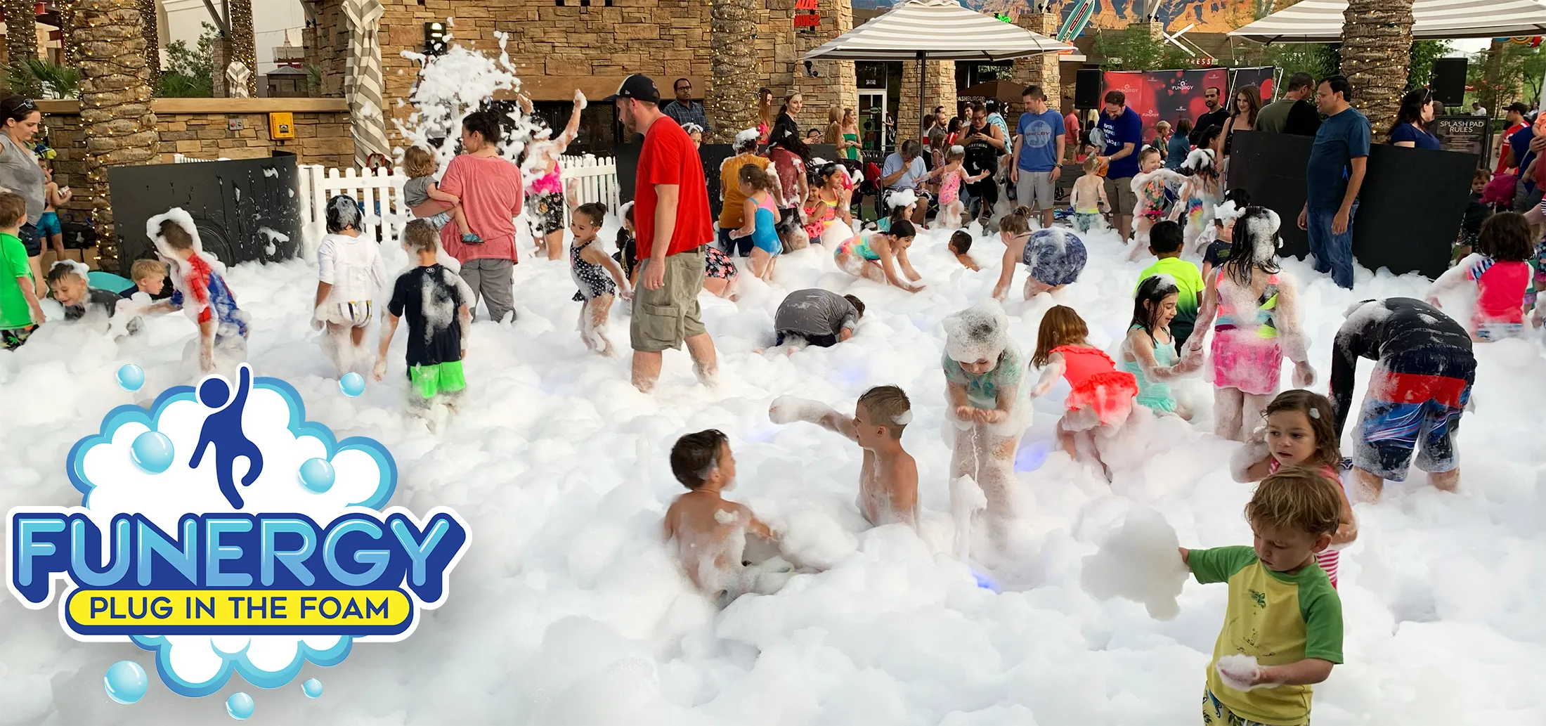 group-event-wide Children and adults play in thick, white foam at an outdoor event. Many kids are covered in bubbles, running and laughing. Adults supervise nearby. Umbrellas and tents can be seen in the background. Funergy logo appears in the corner.