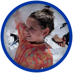 orange-girl A young girl with wet hair, wearing an orange swimsuit with a fruit pattern, smiles while playing in a large amount of foam bubbles. The image has a blue circular border.