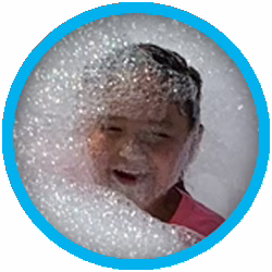 A child wearing a pink shirt is smiling and surrounded by a large amount of foam or bubbles, with their face partially covered.