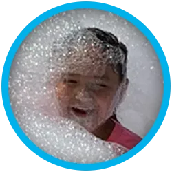 smiling-boy A child wearing a pink shirt is smiling and surrounded by a large amount of foam or bubbles, with their face partially covered.