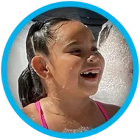 A young girl with dark hair in pigtails, wearing a pink swimsuit, smiles and laughs while playing in foamy bubbles, framed by a blue circular border.