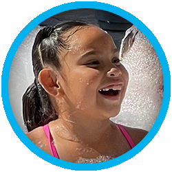 A young girl with dark hair in pigtails, wearing a pink swimsuit, smiles and laughs while playing in foamy bubbles, framed by a blue circular border.