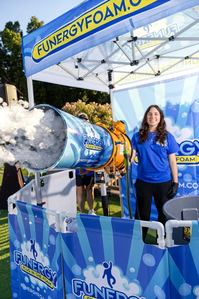 cannon-staffer A smiling woman in a blue shirt stands next to a foam cannon shooting bubbles at an outdoor event under a canopy that says Funergy Foam. The scene is bright and cheerful, with foam and fun-themed decorations.