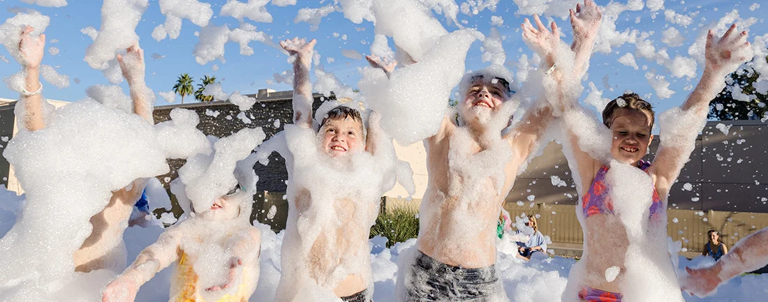 A group of children play joyfully outside, surrounded by a large amount of foam, with their arms raised and smiling faces, under a bright blue sky.