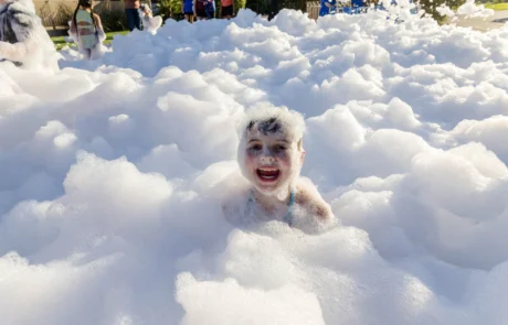 A child with a big smile plays in a large area filled with fluffy white foam, surrounded by other children and sunlight in the background.