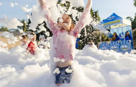 A child playing in thick foam outdoors tosses a large beach ball into the air, smiling joyfully. Other children and a blue event tent are visible in the background under a sunny sky.