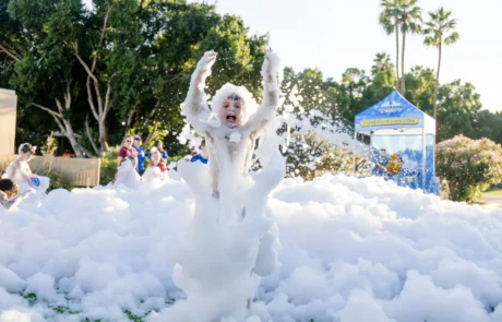 A child covered in white foam raises their arms joyfully in an outdoor area filled with bubbles, with other children playing in the background and trees and a blue tent visible.