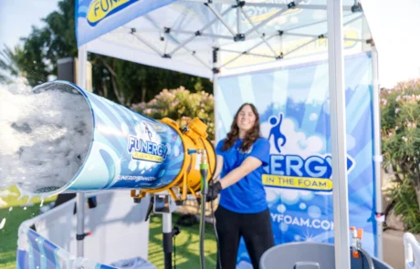 A smiling woman operates a large foam machine under a canopy at an outdoor event, spraying white foam. The machine and canopy feature “FUNERGY IN THE FOAM” branding with blue and yellow colors.