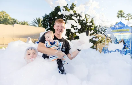 A smiling man holds a baby while standing in thick foam, with a young boy partially submerged beside him. Trees and a tent are visible in the background on a sunny day.