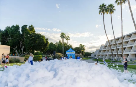Children and adults play in a large pile of white foam outdoors, surrounded by palm trees and buildings on a sunny day, with a blue tent set up in the background.