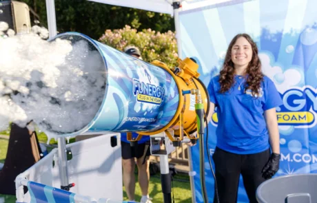 A smiling woman in a blue shirt stands next to a large foam machine at an outdoor event booth decorated with Funergy Foam branding. The machine is releasing a cloud of foam.