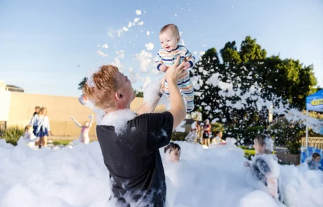 A man joyfully lifts a smiling baby in striped pajamas above his head amid a large area filled with soap bubbles, with children and adults playing in the background outdoors on a sunny day.