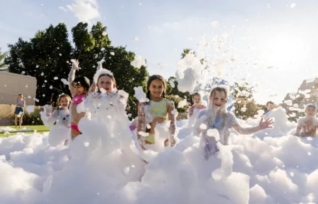 A group of children play joyfully in thick white foam outdoors on a sunny day, smiling and laughing as they are surrounded by bubbles and spray.