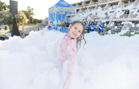 A smiling child stands in thick white foam at an outdoor event, wearing a light pink outfit. People and a blue tent are visible in the background on a sunny day.