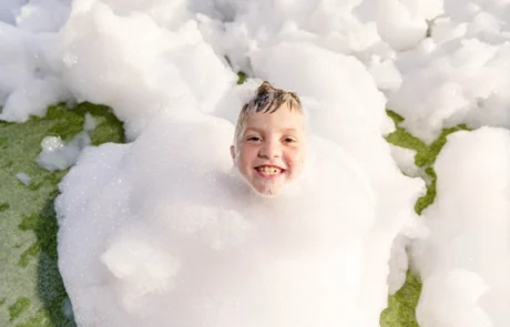 A smiling child’s head pokes out from a large pile of white foam bubbles, with green grass visible beneath the bubbles in the background.