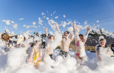 A group of children play joyfully in a large area filled with foam, throwing bubbles in the air under a clear blue sky. They are smiling and wearing swimsuits, enjoying the outdoor activity.