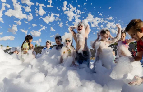 A group of children play joyfully in thick, white foam outdoors under a bright blue sky. Some kids laugh and raise their arms while foam bubbles float in the air around them.