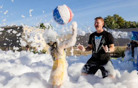 A young girl and a man play with a beach ball in a large area filled with foam outdoors. The man is wearing a tuxedo-print shirt, and both are covered in bubbles, enjoying the sunny day.