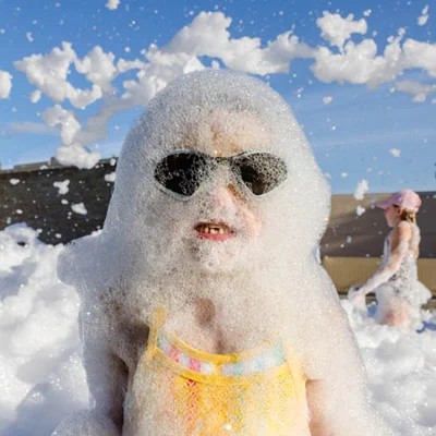 Goggles Are Recommended A child wearing sunglasses and a swimsuit is covered in bubbles, standing outdoors on a sunny day. Another child in a hat is visible in the background, also surrounded by foam and bubbles.