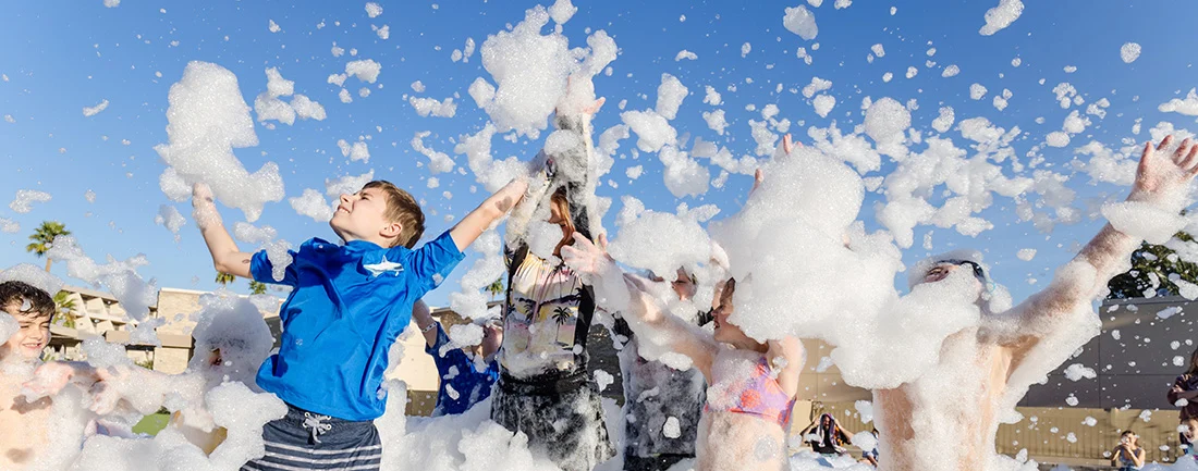 Children joyfully play in a large cloud of foam outdoors on a sunny day, with arms raised and bubbles flying in the air. The sky is clear and blue in the background.