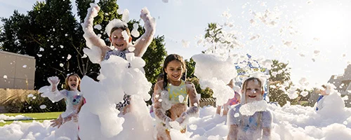 service-area-wide-1 Children joyfully play in a large area filled with white foam outdoors on a sunny day. They are laughing and covered in bubbles, with trees and buildings visible in the background.