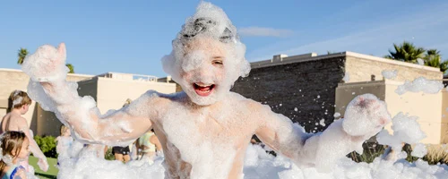 service-area-wide-2 A child covered in soap bubbles smiles and raises their arms while playing at an outdoor foam party on a sunny day. Other children and buildings are visible in the background.