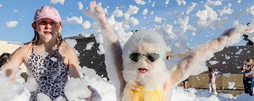 service-area-wide-3 Two children play in a large pile of foam outdoors. One child wears sunglasses and is covered in bubbles with arms raised, while the other smiles and wears a pink cap. The sky is clear and blue.