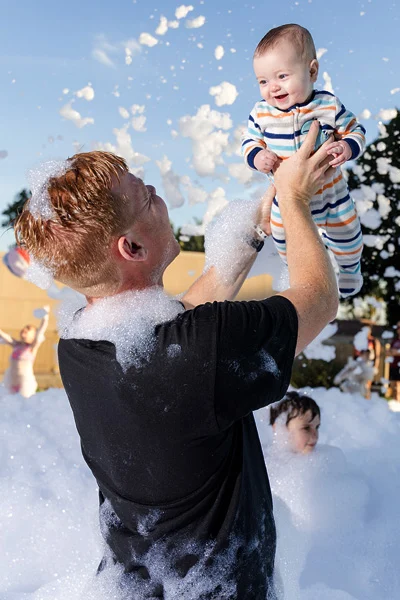 vertical-baby-dad A man covered in foam lifts a smiling baby in striped pajamas above his head at an outdoor foam party, with bubbles floating around and another child in the background.