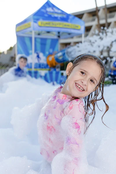 vertical-girl-smiling A smiling young girl in a pink swimsuit plays in a large area filled with foam, with another child and a foam machine visible in the background under a blue tent outdoors.